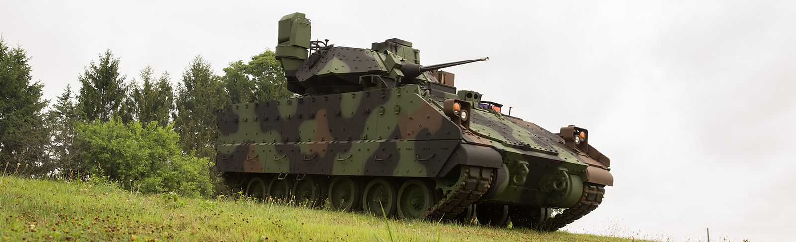 A green camouflage Bradley vehicle driving down a grassy hill