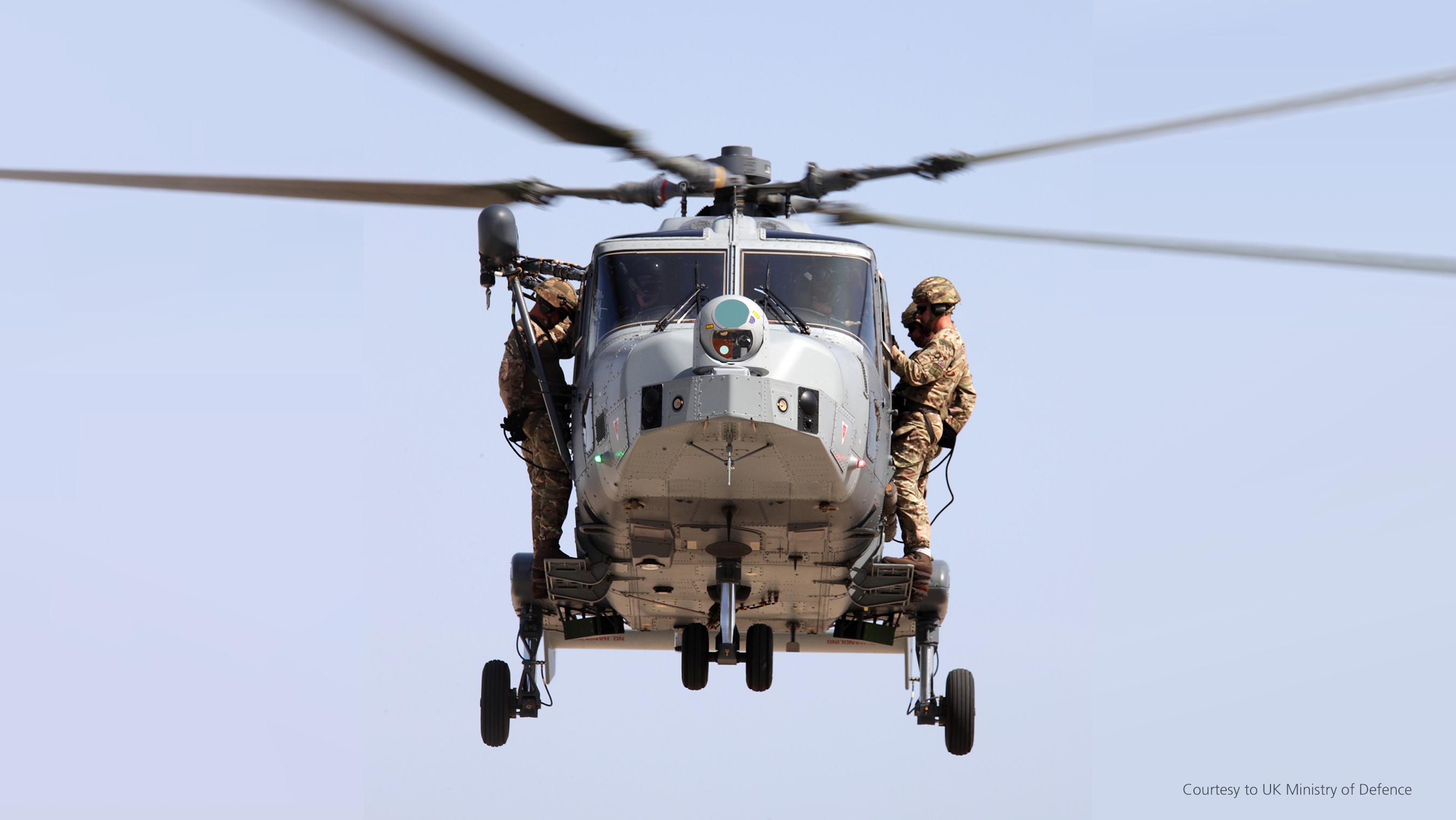 Wildcat rotary-wing aircraft midflight against clear blue sky with four soldiers riding on the exterior, two standing on either side. Faint view of the pilot and co-pilot seated in their seats inside the chopper.