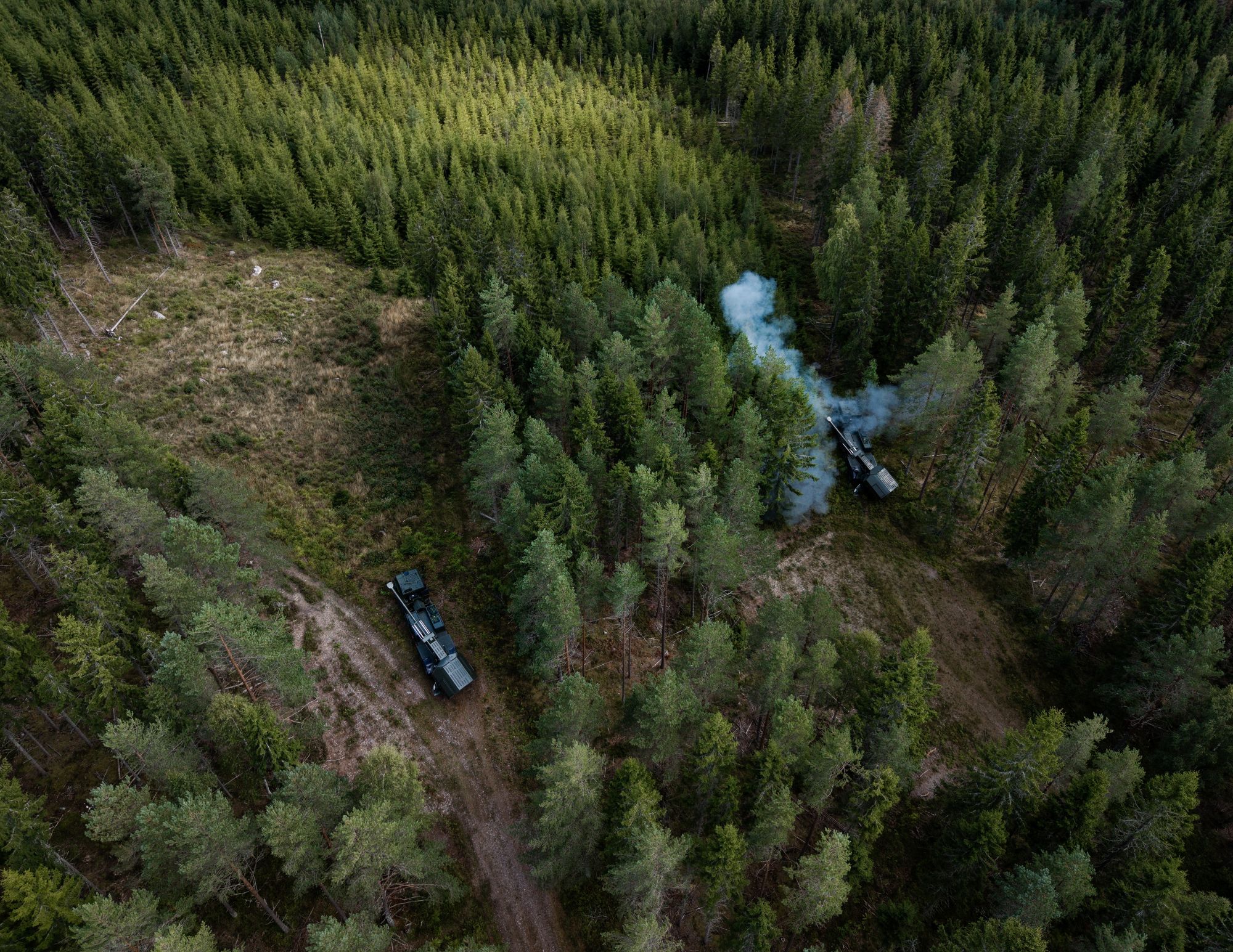 Drone footage captures two ARCHER 8x8 self-propelled artillery systems in a forested area, with one vehicle firing a live round during a field exercise.