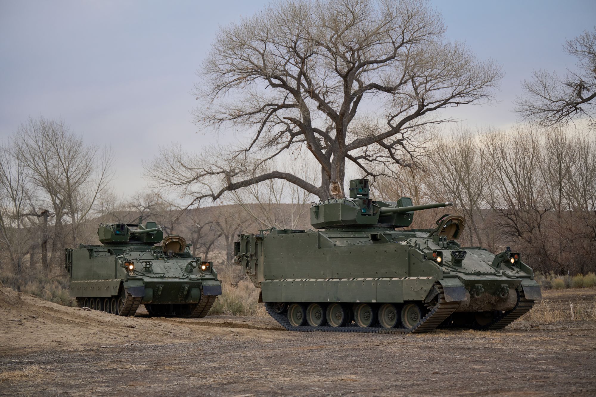 Two green tracked combat vehicles drive through mountainous terrain.