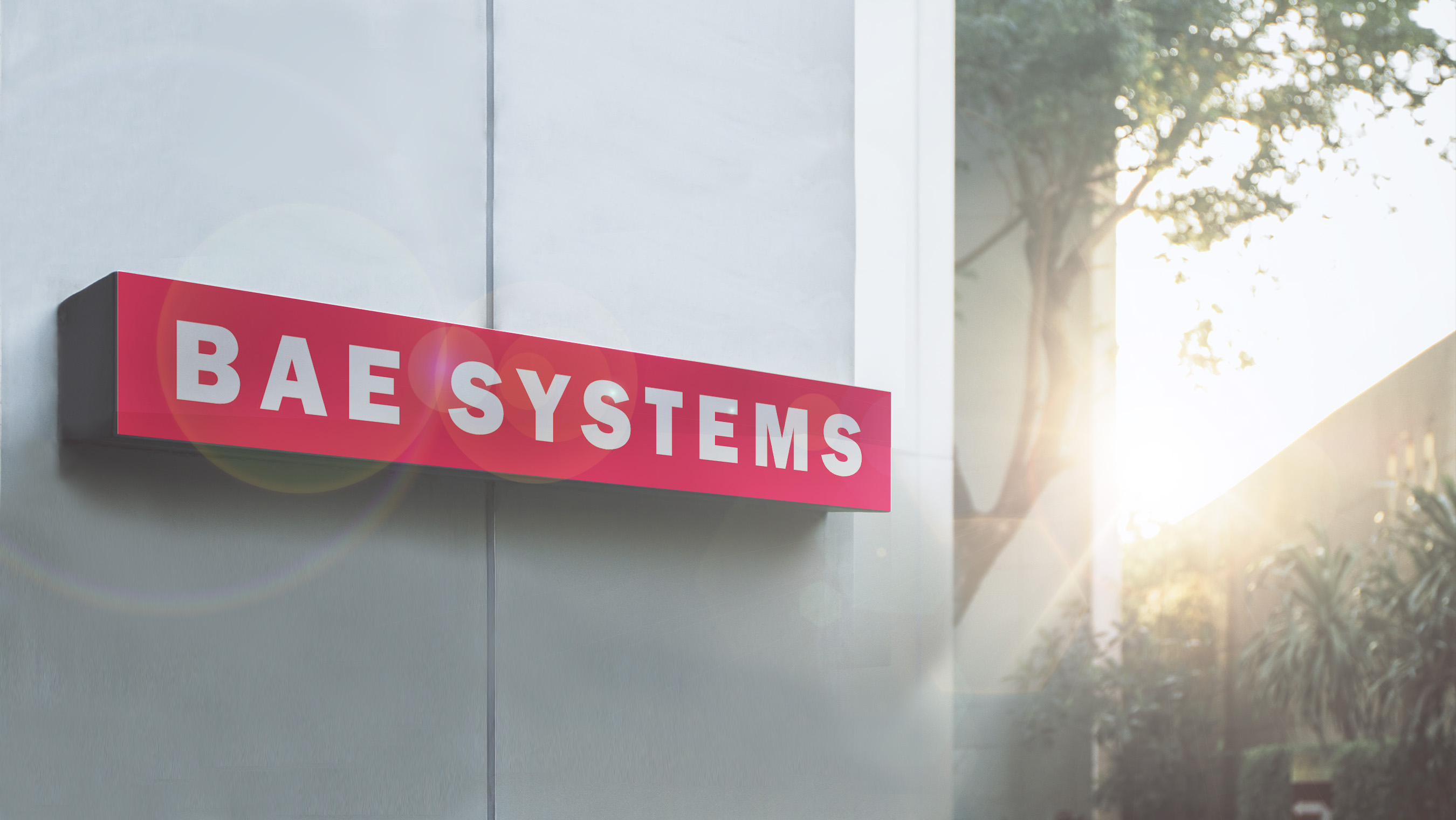  BAE Systems sign on corner of non-descript gray building with trees and sunlight in background 