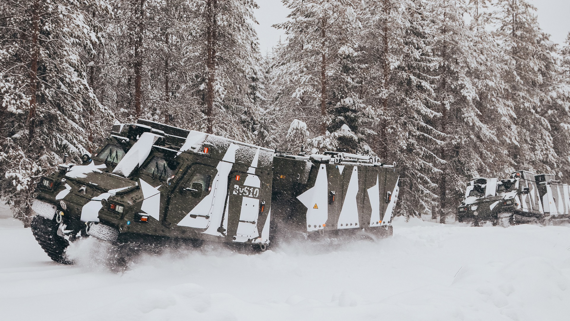 A white and gray camouflage BvS10 drives through snow with snow-covered trees behind it 