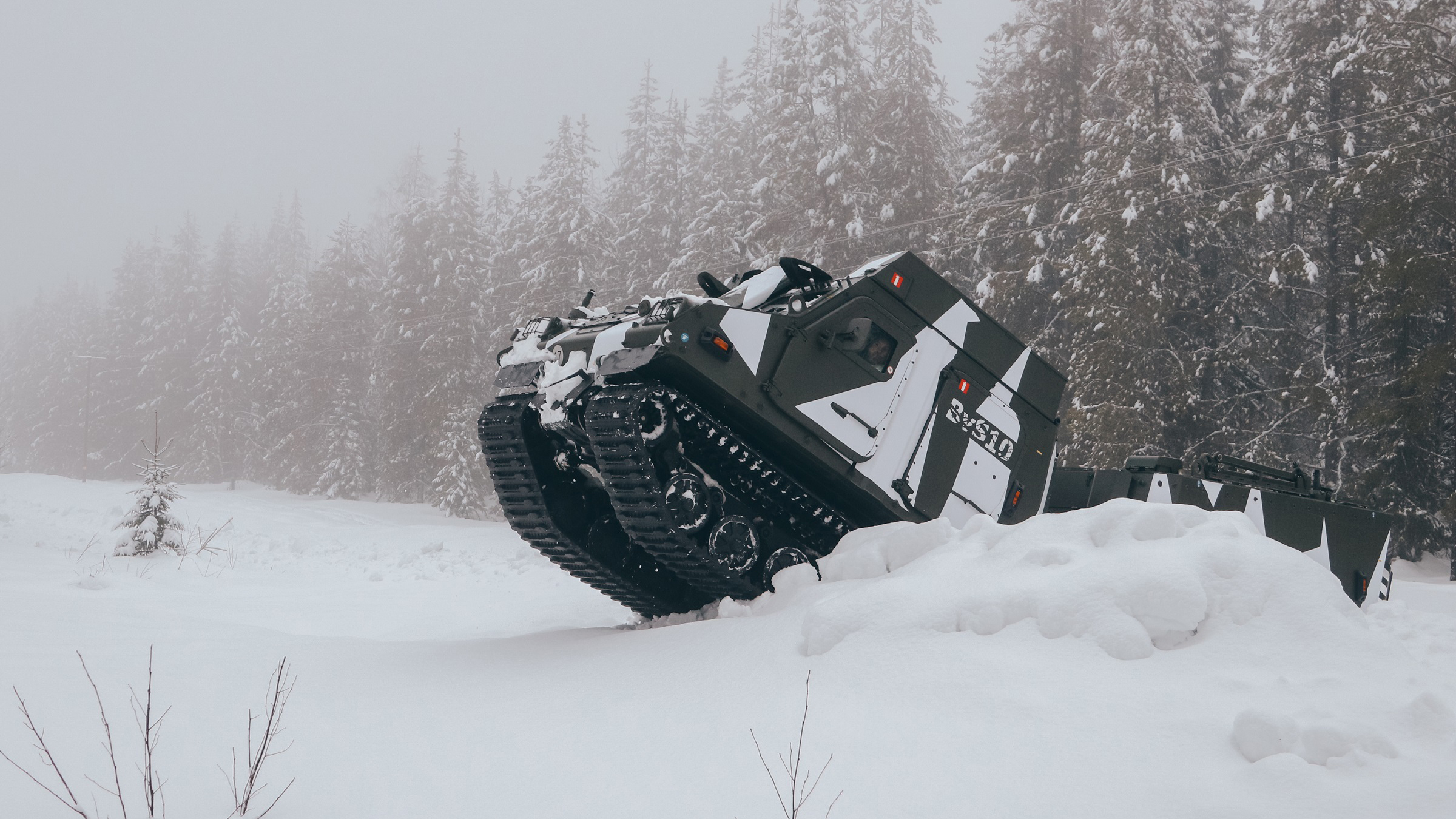 A gray and white camouflage BvS10 goes over a snow hill with snow covered trees in the background 