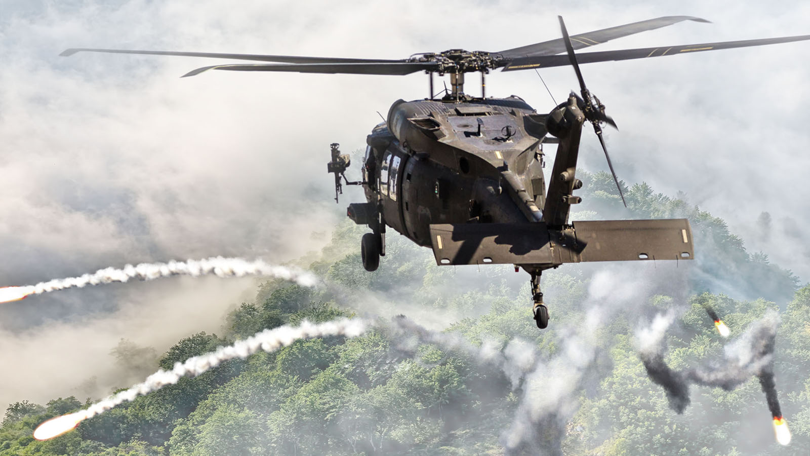 A Black Hawk helicopter fires countermeasure flares. Fog hovers over a dense forest below.