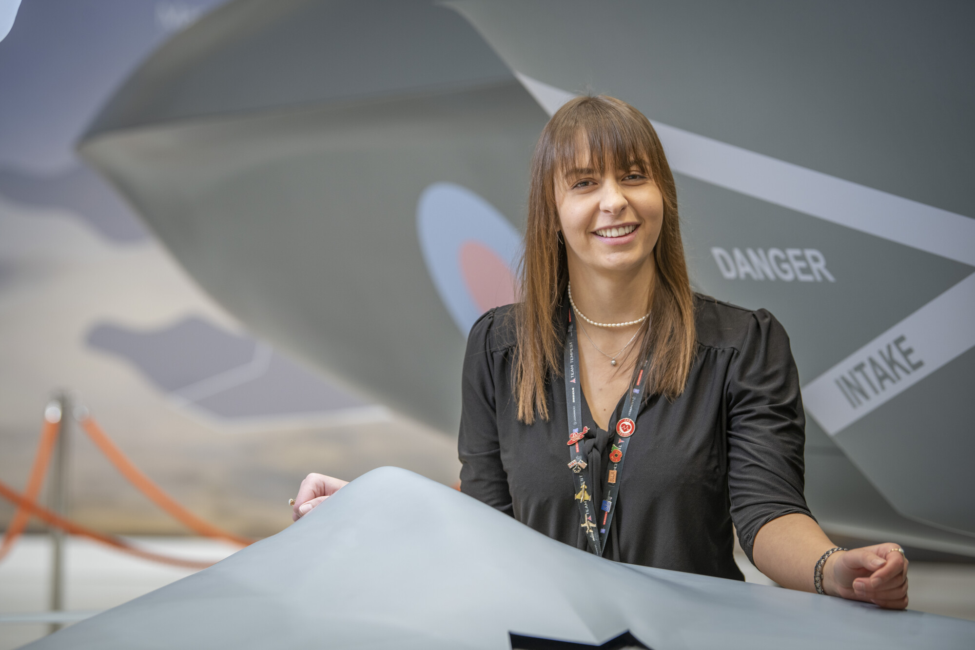 Amy smiling at the camera standing in a hangar in front of a small Tempest concept model with the full scale model behind