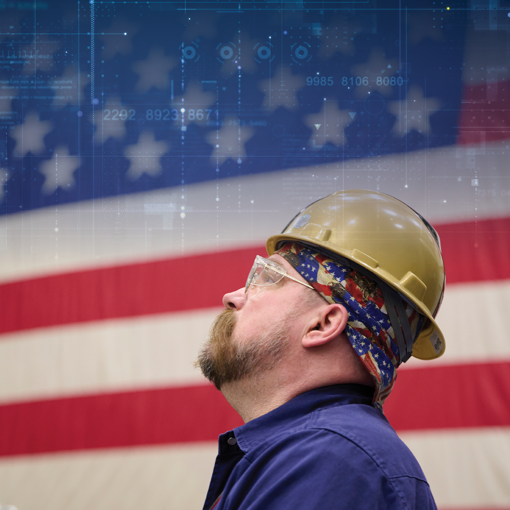 A BAE Systems employee in a hard hat looks up with an American flag in the background
