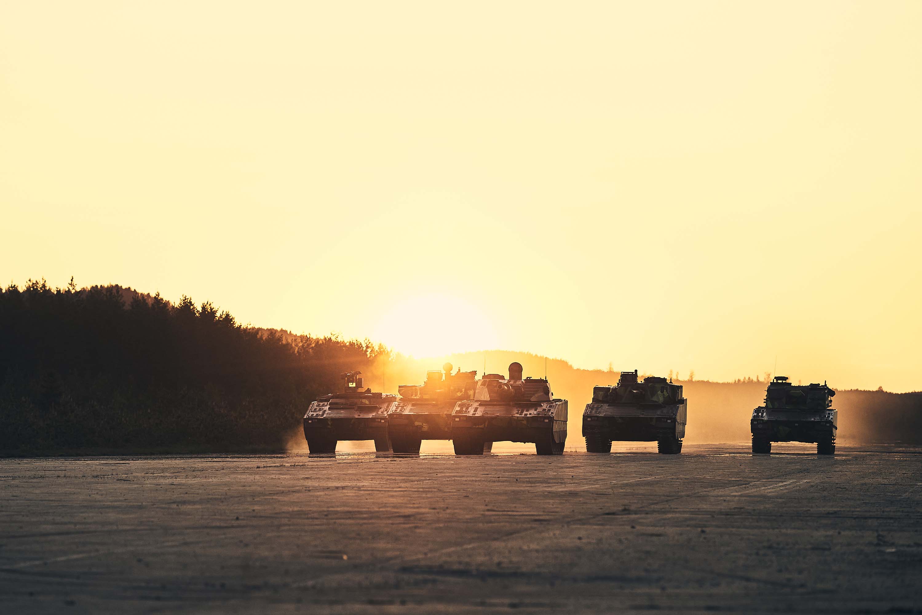 5 CV90s driving down the road as the sun sets behind them