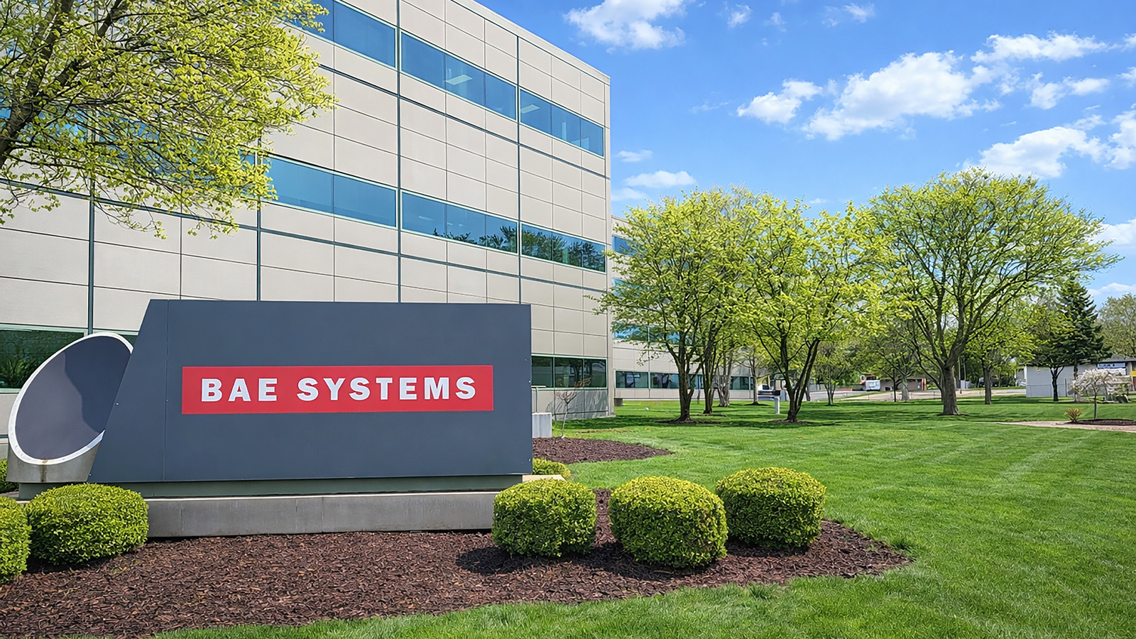 Exterior of building with BAE Systems signage in front, green grass and trees, cloudy sky in background 