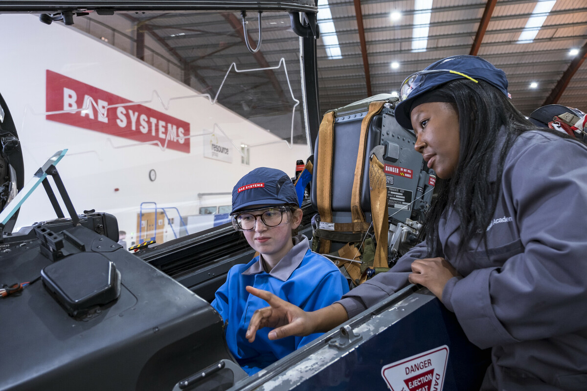 An image of two workers wearing work clothing. One is stitting in a cockpit listening. One of them is talking and showing the other the cockpit.
