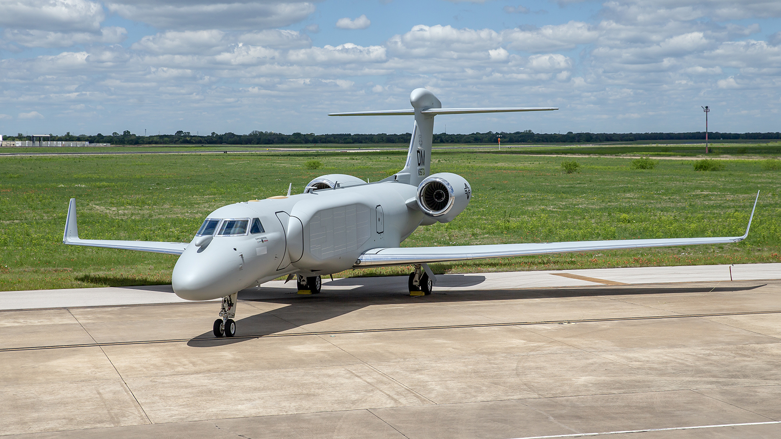 A photo shows an EA-37B parked at an airfield, with green grass and a cloudy blue sky behind it. 