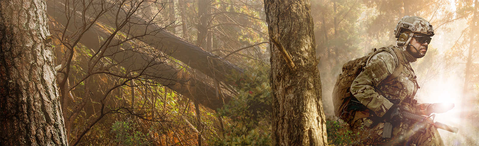 Soldier wearing camo gear carrying rifle and backpack traveling through dense forest with sunshine rays in background. 