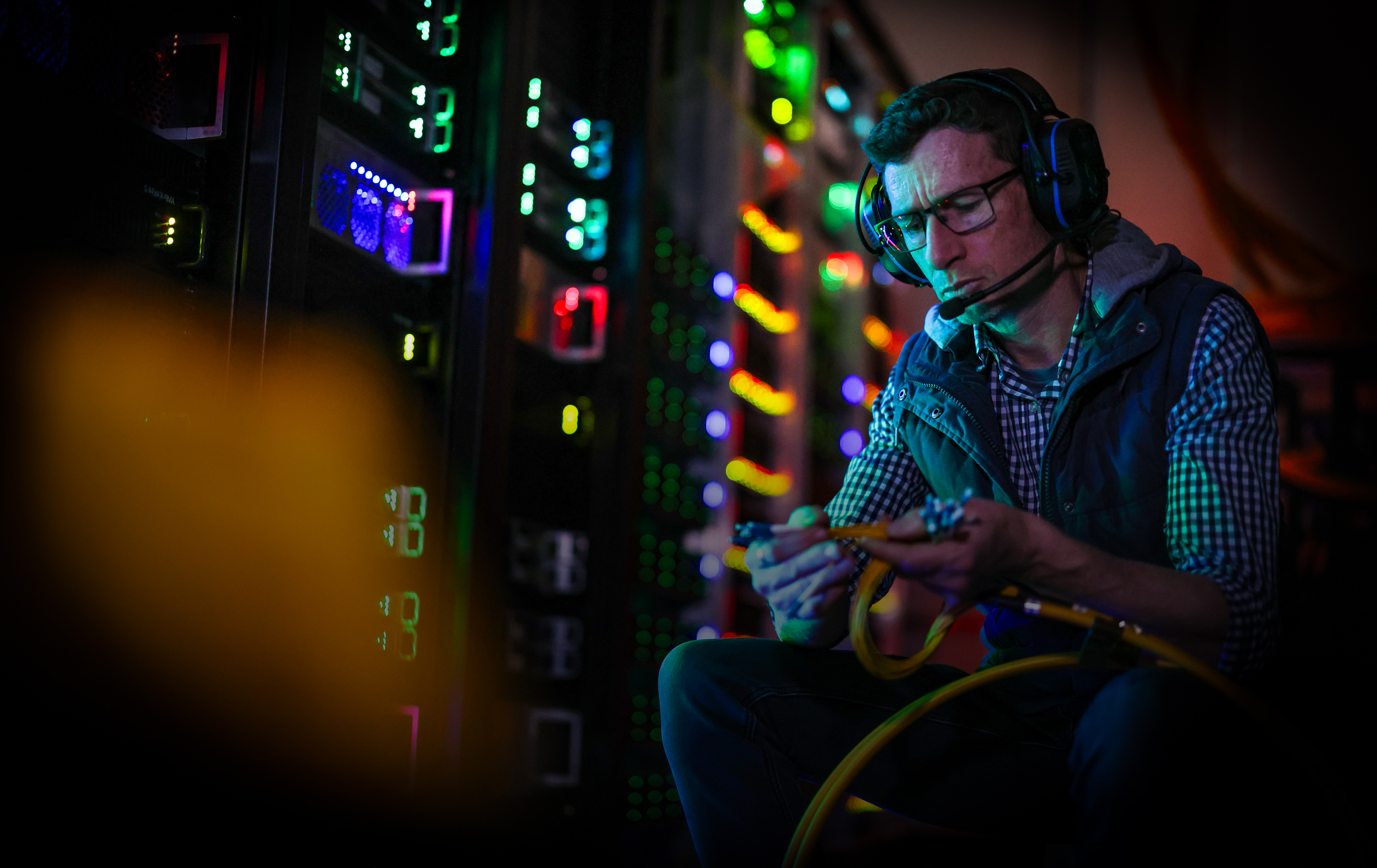 Person wearing a communications headset connecting network cables in a server room beside illuminated equipment racks.
