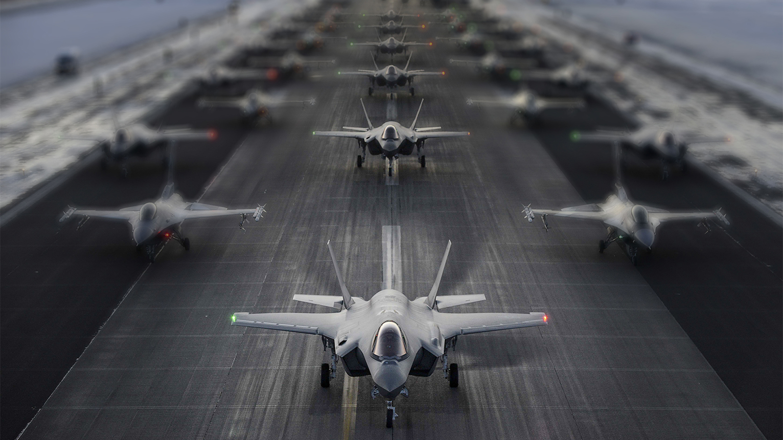 A column of F-35s line a runway, flanked by F-16s.