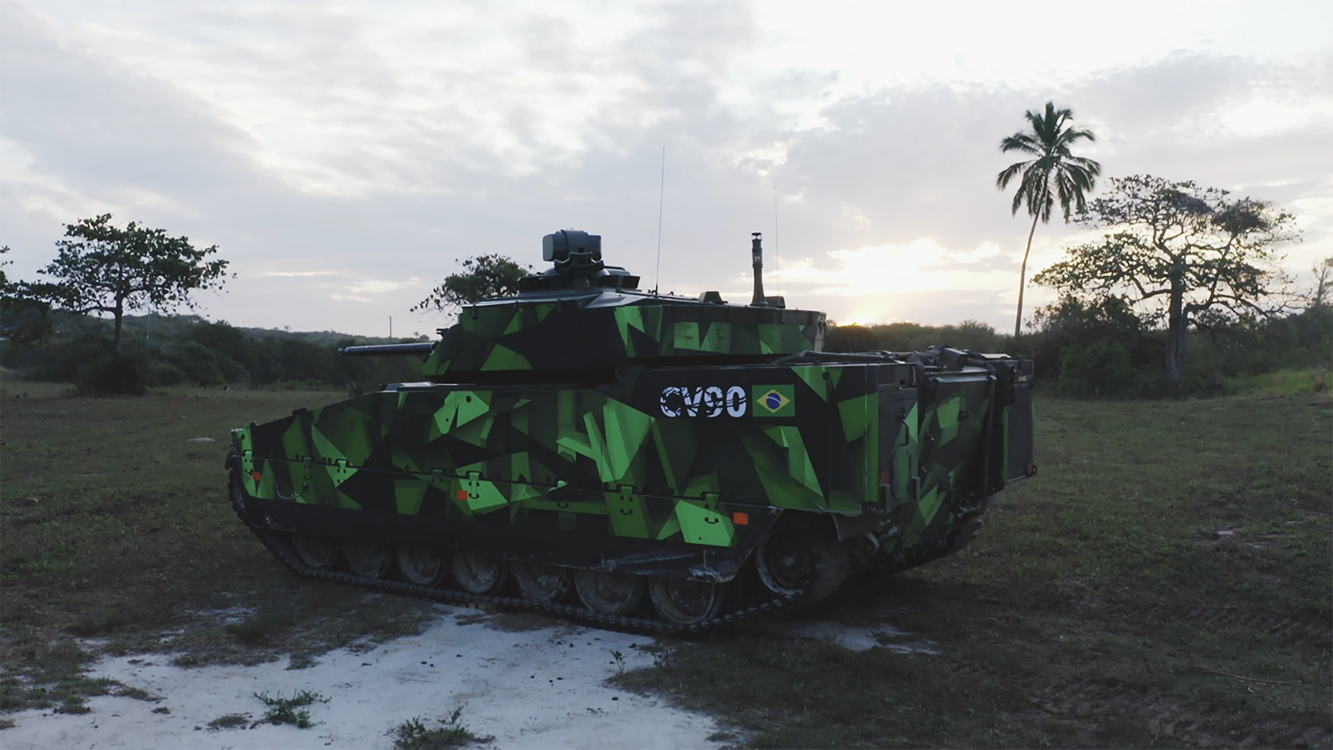 A green CV90, with "CV90" and the Brazilian flag on it sits in a field with sandy patches