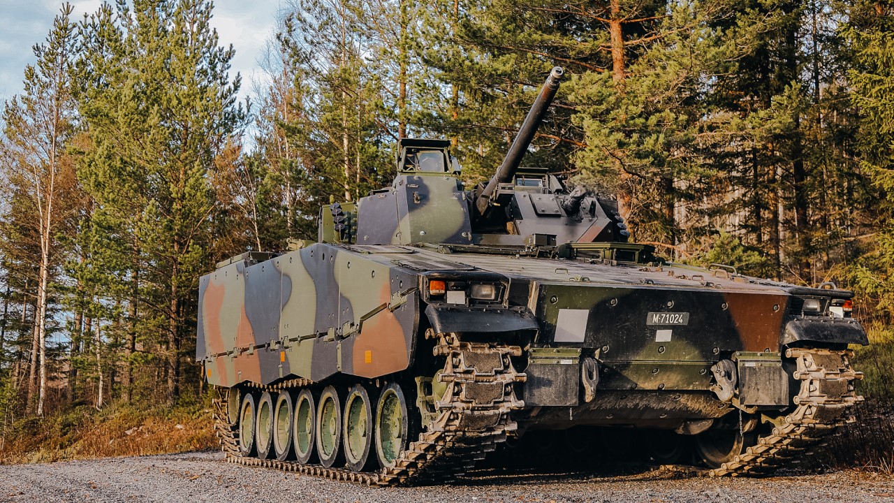 BAE Systems Hagglunds' CV9030 in camo paint positioned on a gravel road next to an evergreen forest. 