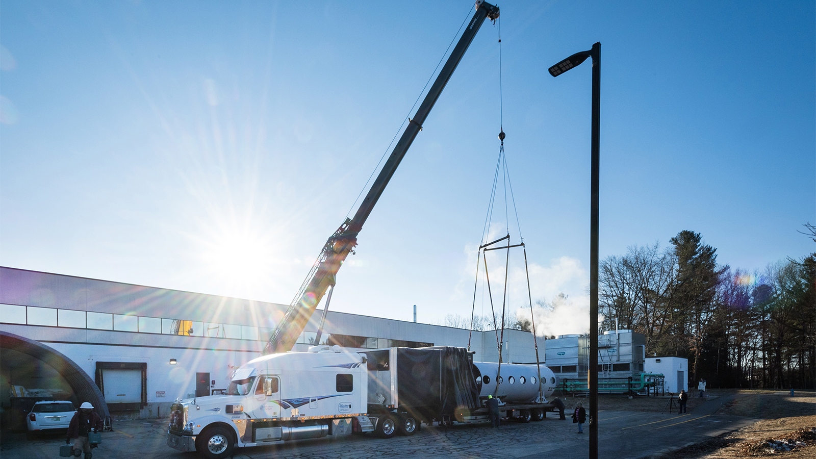 Large crane removing fuselage from flatbed of white truck outside white BAE Systems building with sun streams in background