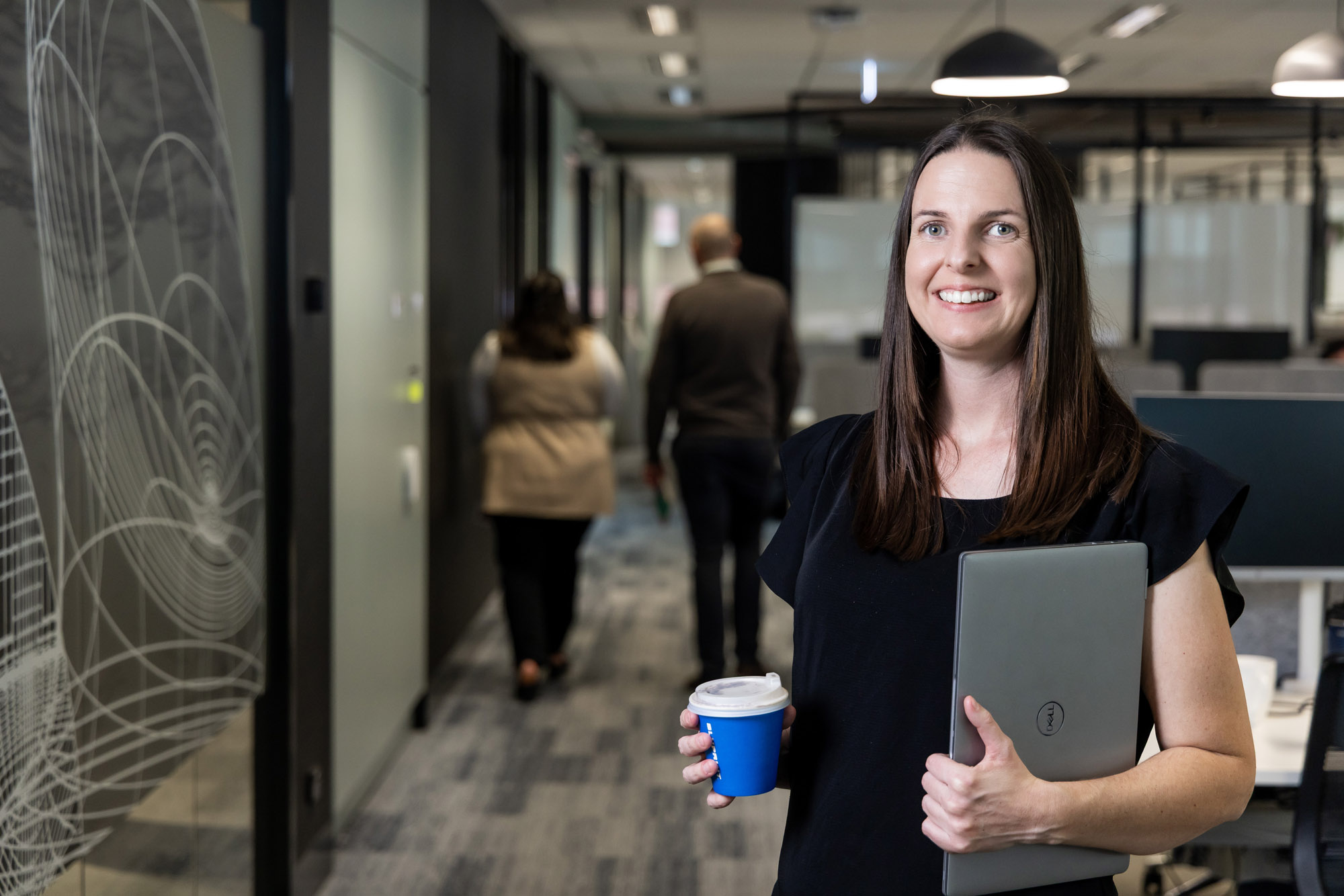 Brenna holding a computer and coffee, standing in an office environment, smiling and looking at the camera