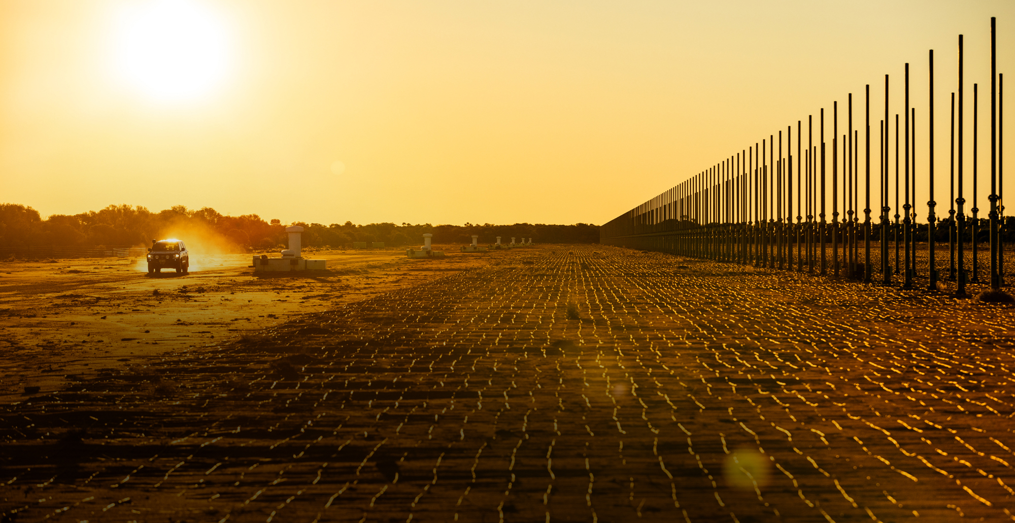 Photograph of JORN radar site in the late afternoon, with a low, hot sun beaming down on the radar. A 4wd vehicle drives along the dusty ground parallel to the radar equipment.