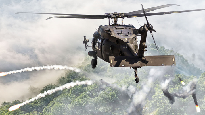A Black Hawk helicopter fires countermeasure flares. Fog hovers over a dense forest below. A Black Hawk helicopter fires countermeasure flares. Fog hovers over a dense forest below.